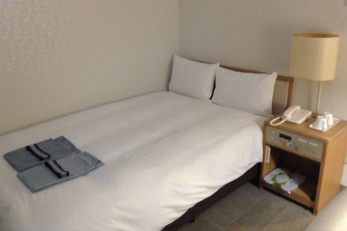 A hotel room with a single bed, a bedside table with a lamp, a phone, and a small drawer with a tissue box.