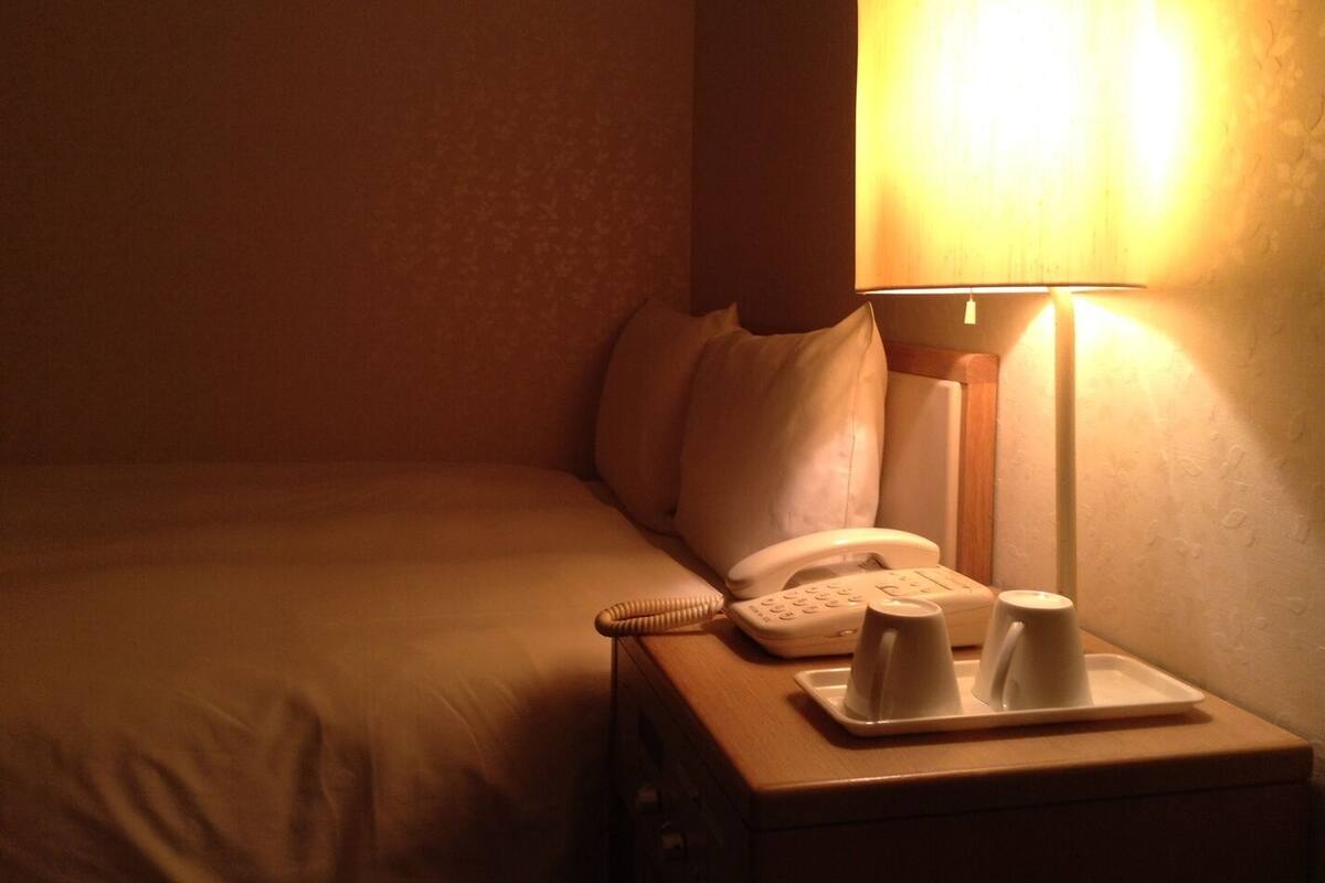 A hotel room with a bed, a bedside table with a lamp, a telephone, and a tray with cups.