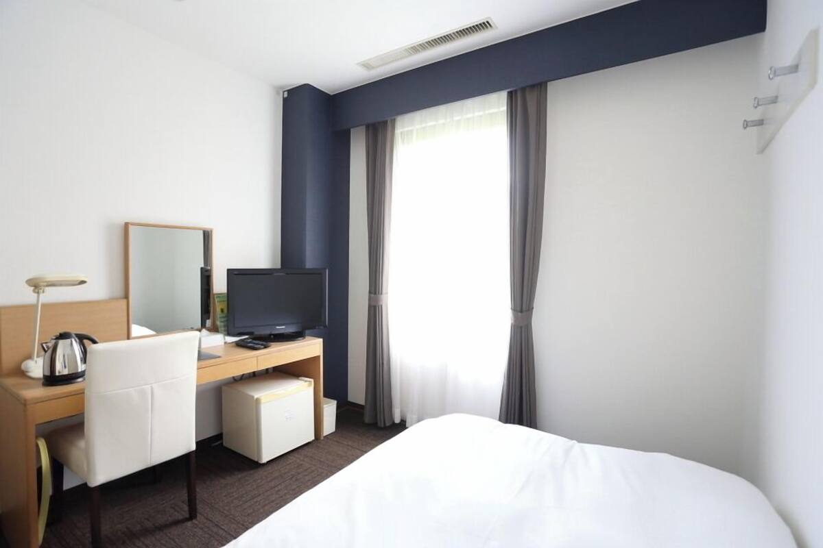 A hotel room with a bed, a desk with a computer, a chair, a television, and a window with curtains.