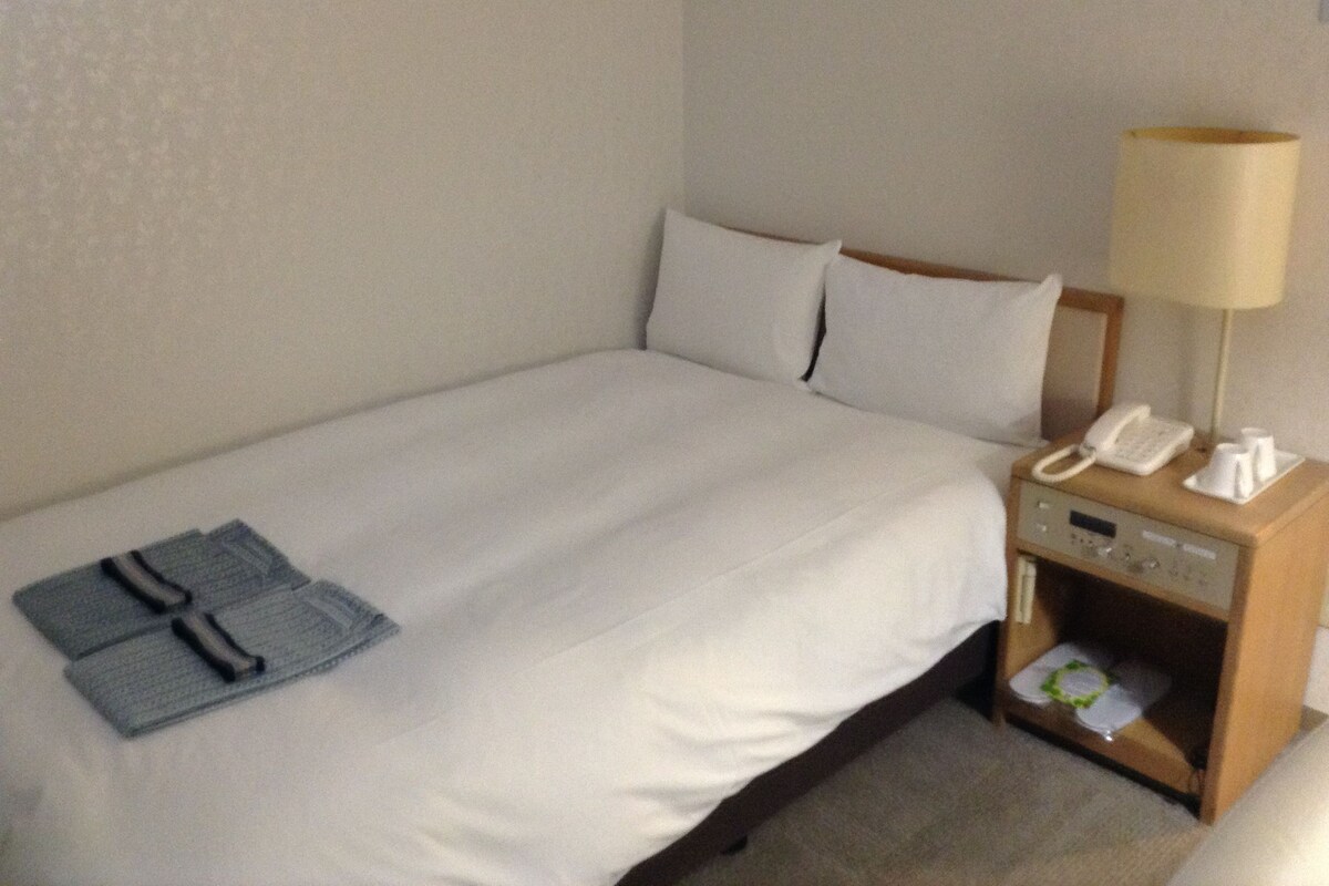 A neatly made bed with white linens, a bedside table with a lamp, a phone, and a small tray with cups.