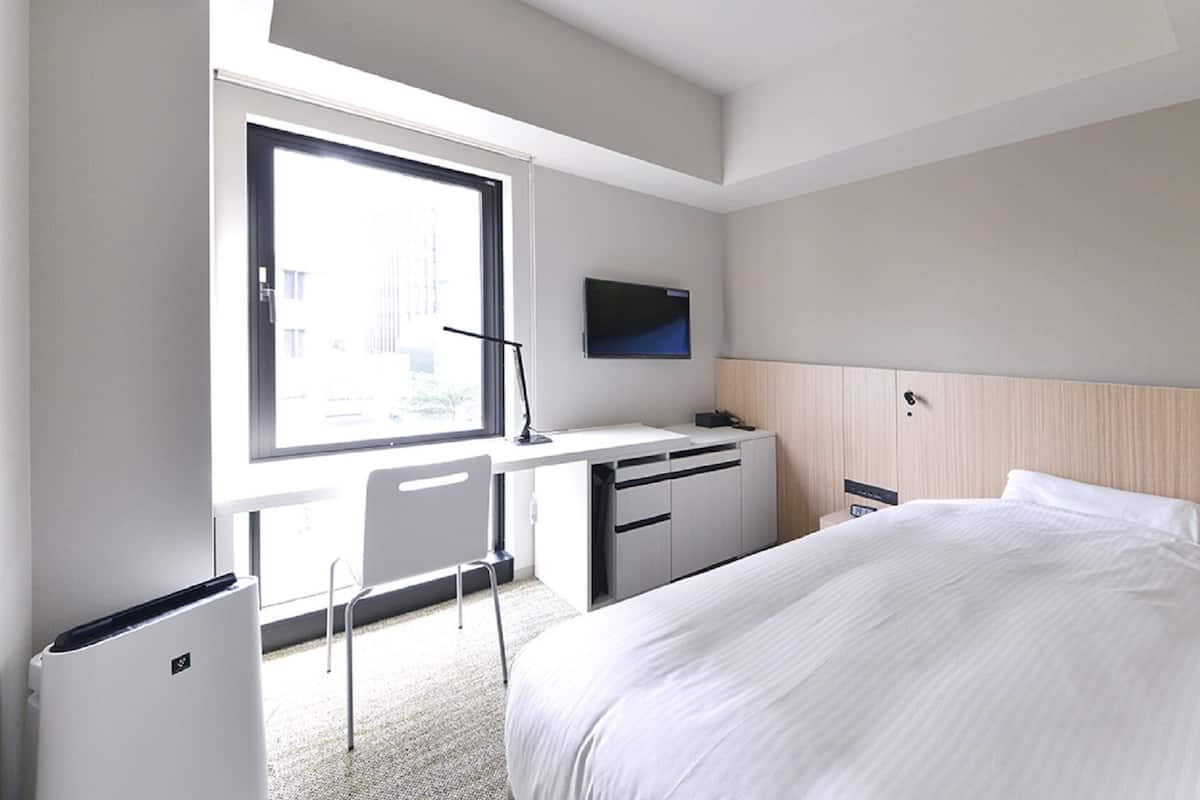 Quarto de hotel com cama, escrivaninha, cadeira, televisão e janela.