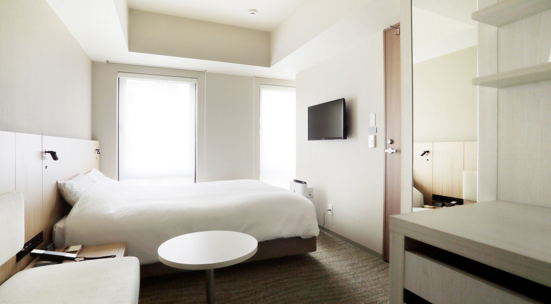 A hotel room with a bed, bedside table, chair, round table, and a TV mounted on the wall.