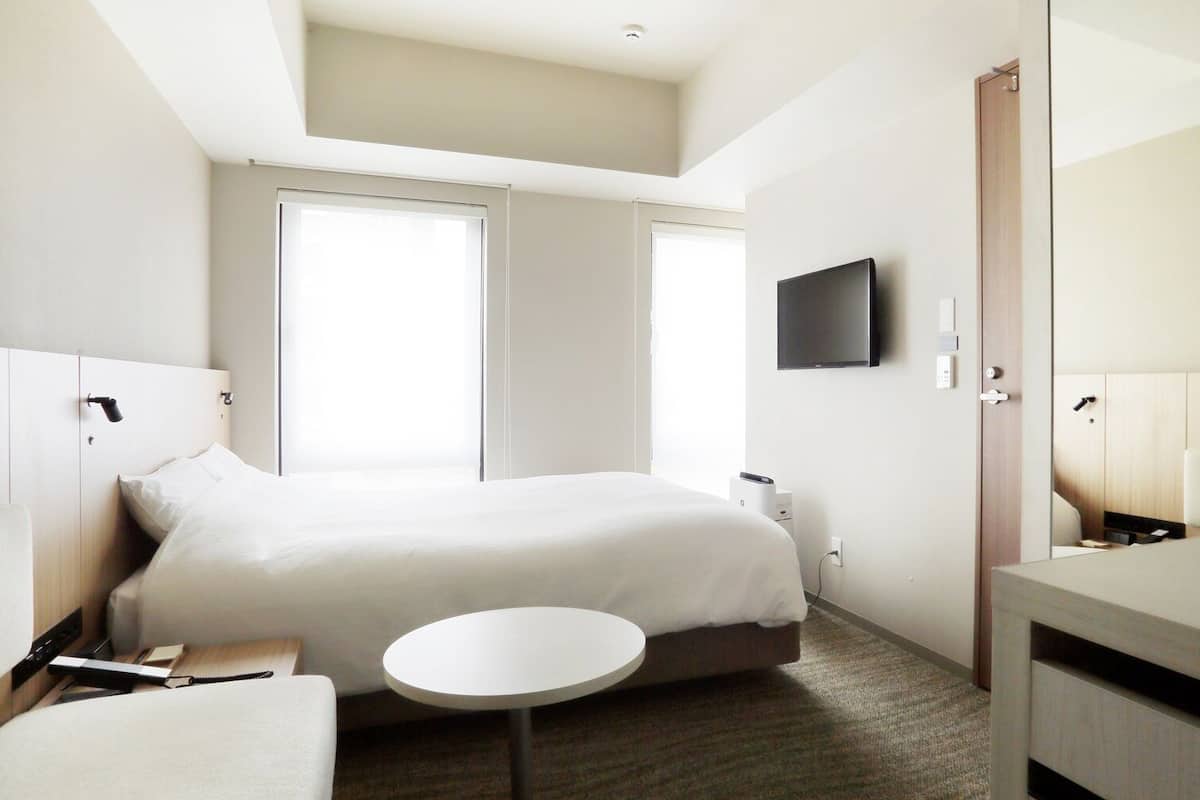 Quarto de hotel com cama, mesa de cabeceira, cadeira, mesa redonda e televisão fixada na parede.