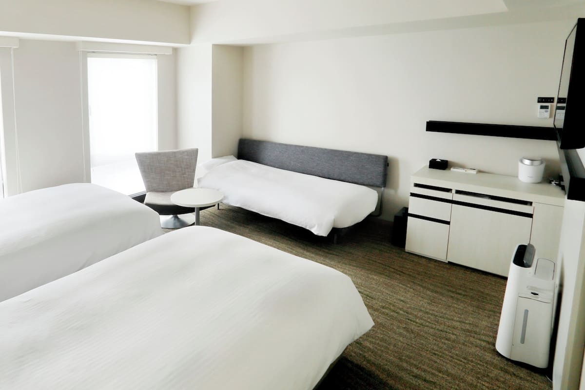 Quarto de hotel com duas camas, uma mesa redonda pequena, uma cadeira, uma escrivaninha com um computador e uma televisão.