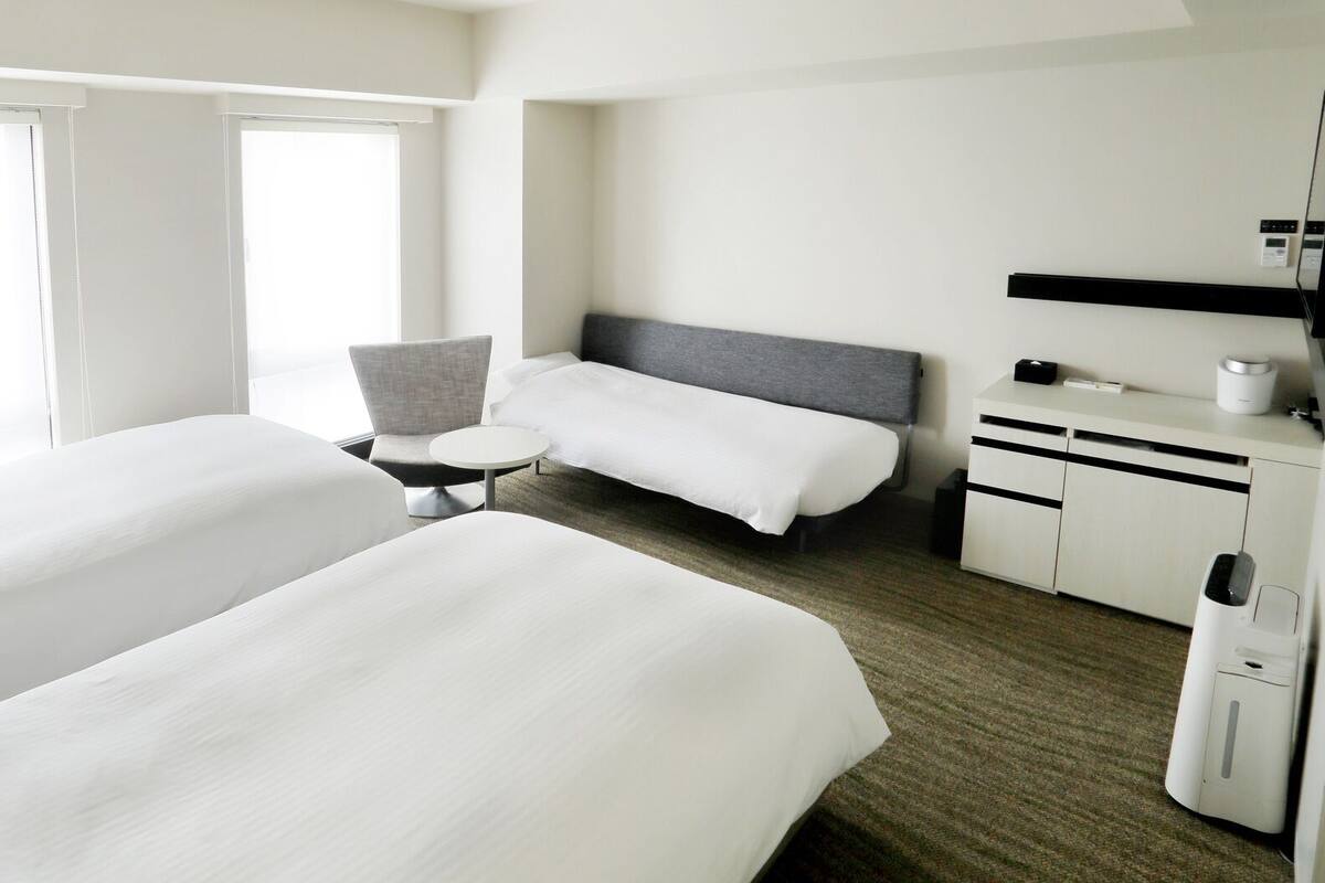 Quarto de hotel com duas camas, uma mesa redonda pequena, uma cadeira, escrivaninha com um computador e uma televisão.