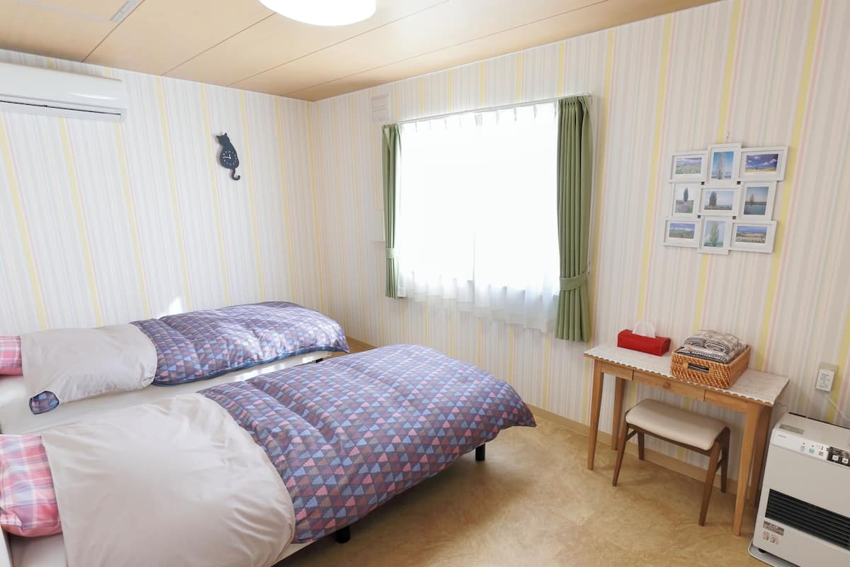 Ein Zimmer mit zwei Betten, einem kleinen Tisch, einer Bank, einer Klimaanlage und einem Fenster mit Vorhängen.