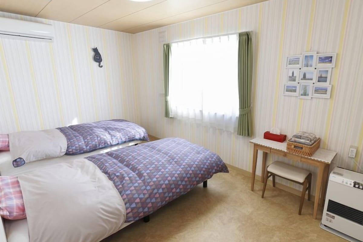 A bedroom with two beds, a small table, a bench, a wall-mounted heater, and a window with curtains.