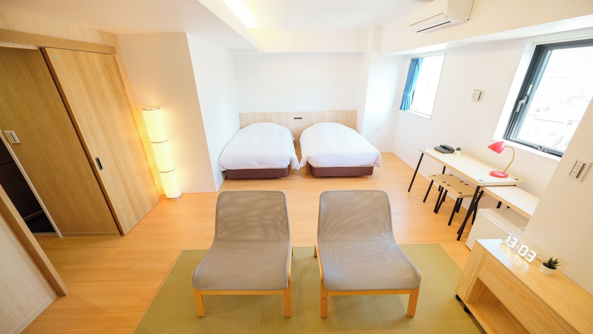 A compact room with two single beds, a desk with a chair, a small table with a lamp, and a window with curtains.