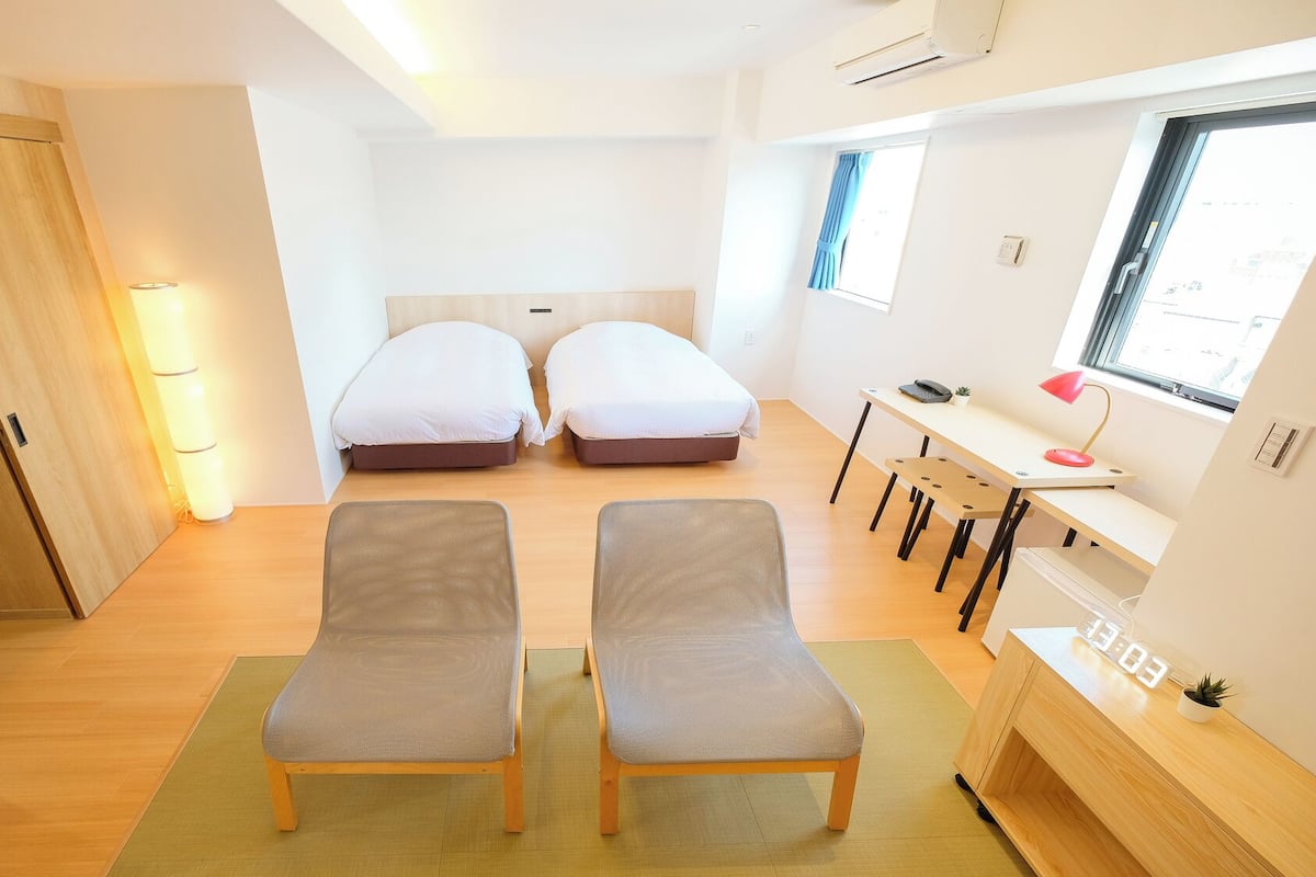 Una habitación pequeña con dos camas individuales, un escritorio con silla, una mesita con lámpara y una ventana con cortinas.