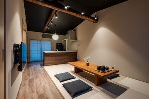 A modern interior with a wooden dining table, black cushions, a hanging light, and a kitchen in the background.