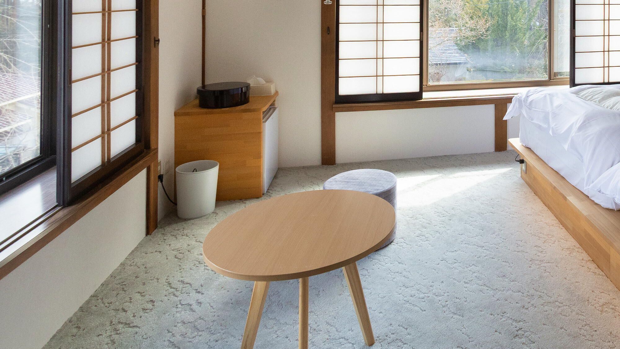 Quarto com piso de tatame, uma mesa de madeira baixa, cama com roupa de cama branca e janela com portas shoji.