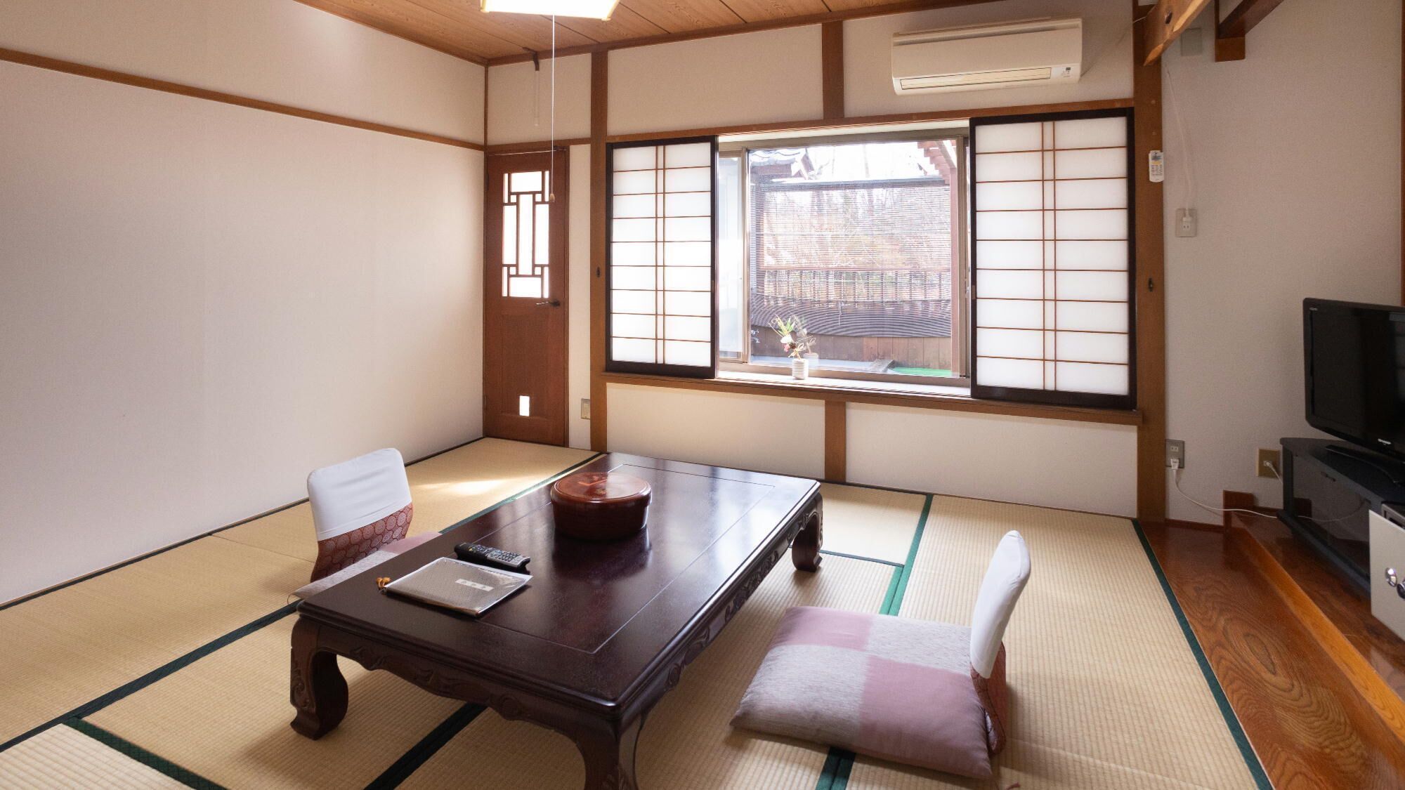 Um quarto japonês tradicional com tatames, uma mesa de madeira baixa, um banco acolchoado e uma porta corrediça com vista para um jardim.