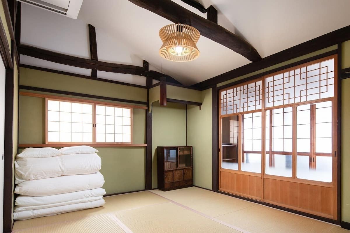 Um quarto japonês tradicional com tatames, futon, portas deslizantes e luminária pendente.