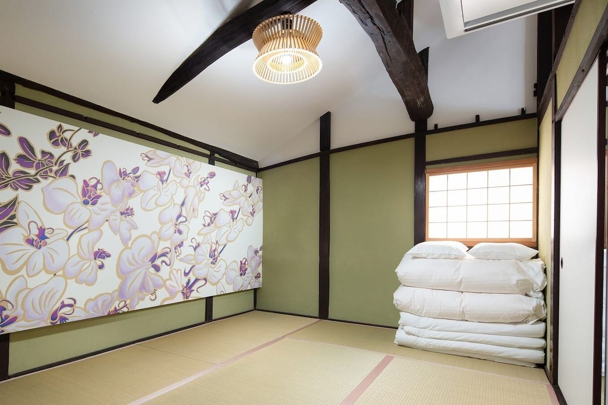 Um quarto japonês tradicional com piso de tatame, um painel floral na parede e uma pilha de roupa de cama branca.