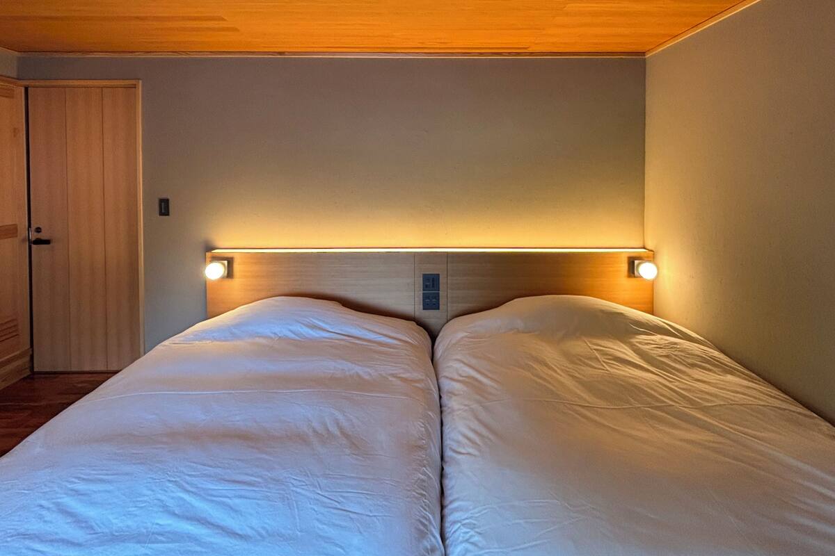 Cama doble con dos lámparas de noche, techo de madera y una puerta a la izquierda.