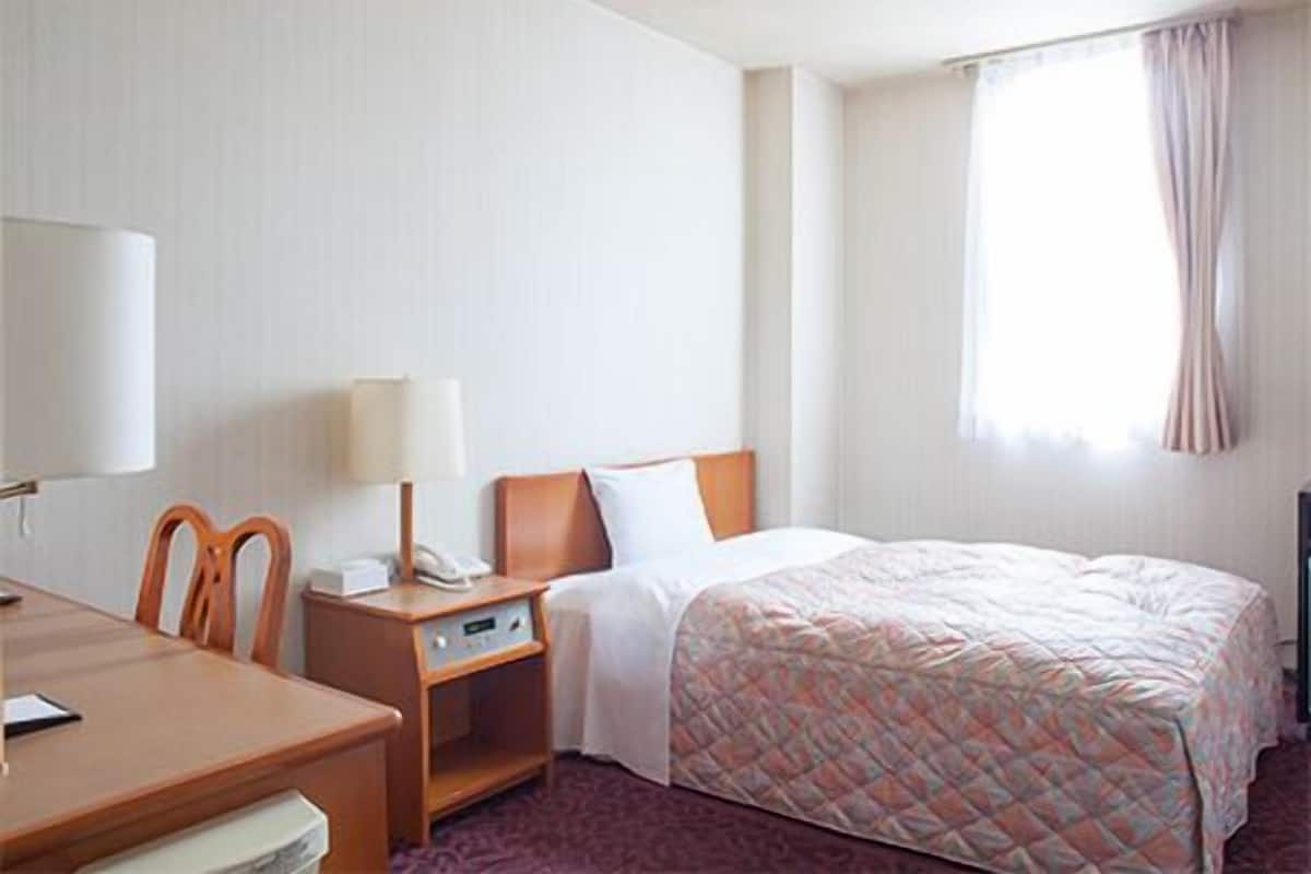 Una habitación de hotel con cama, escritorio con silla, teléfono y ventana con cortinas.