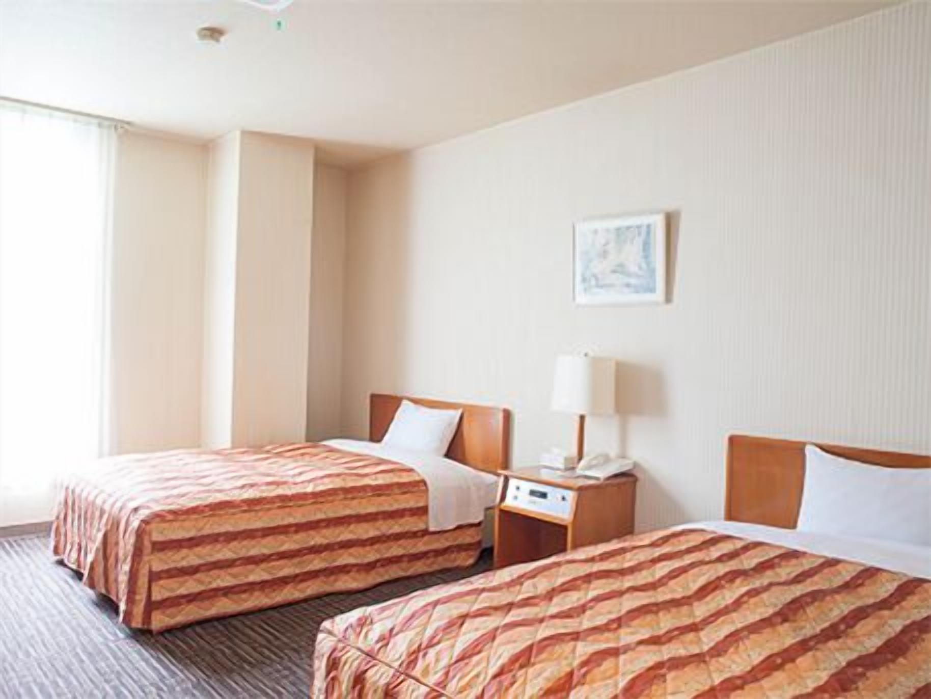 A hotel room with two beds, a bedside table with a lamp, and a window.
