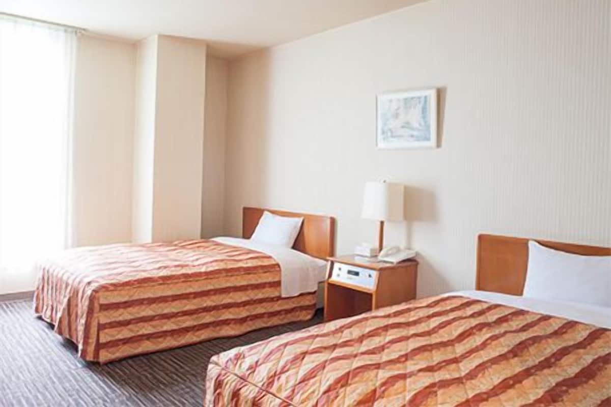 Una habitación de hotel con dos camas, una mesita de noche con lámpara y una ventana.