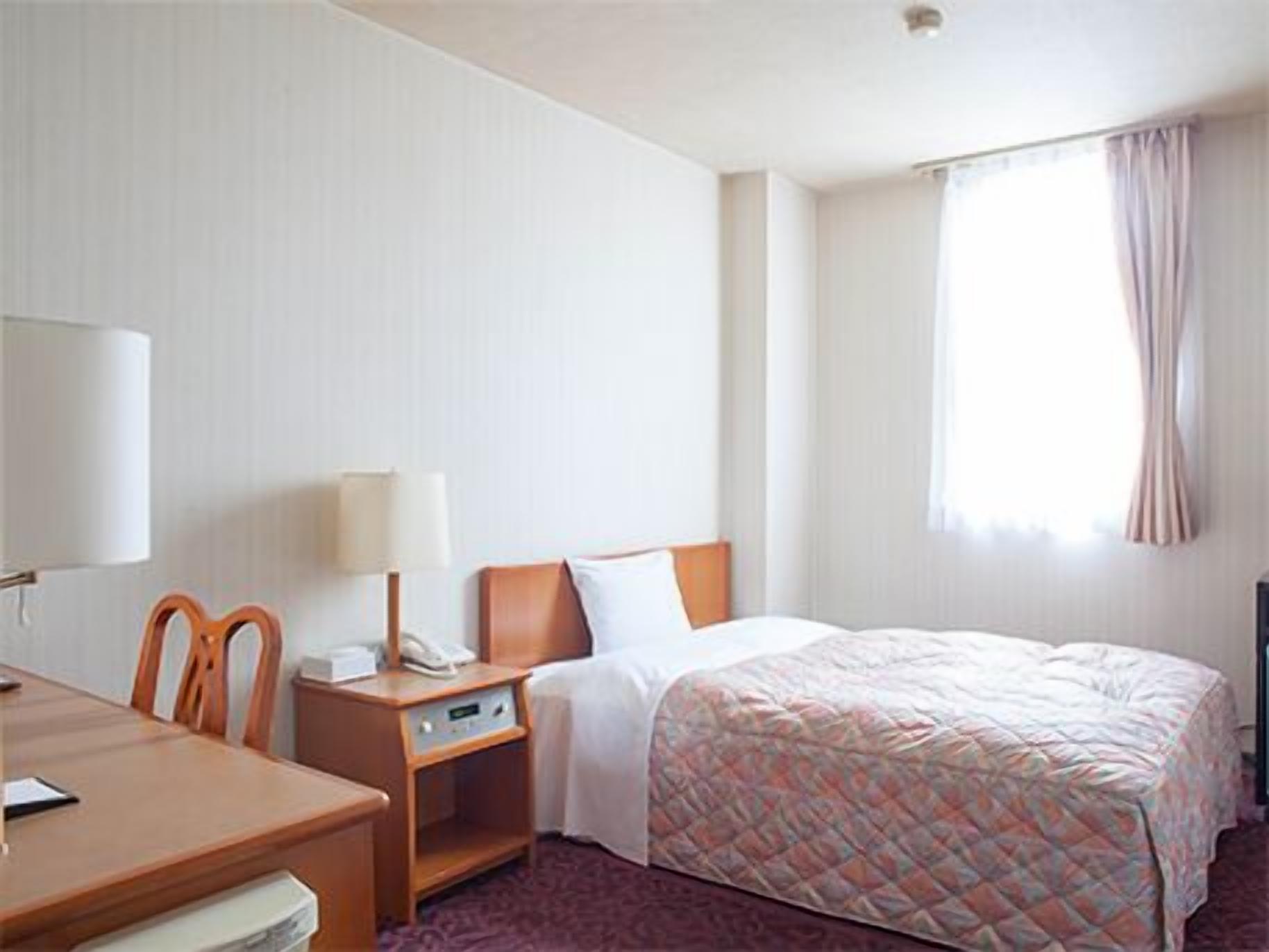 A hotel room with a bed, a desk with a lamp, a chair, and a telephone.