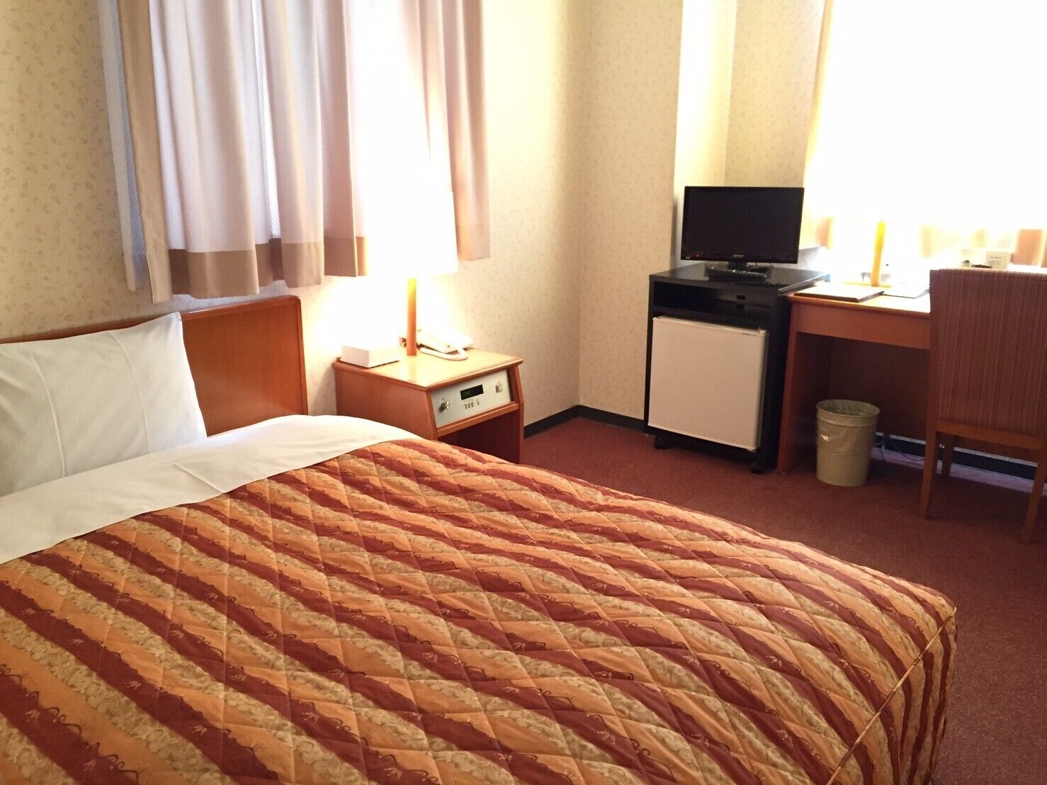 A hotel room with a bed, a desk, a chair, a trash bin, and a television.
