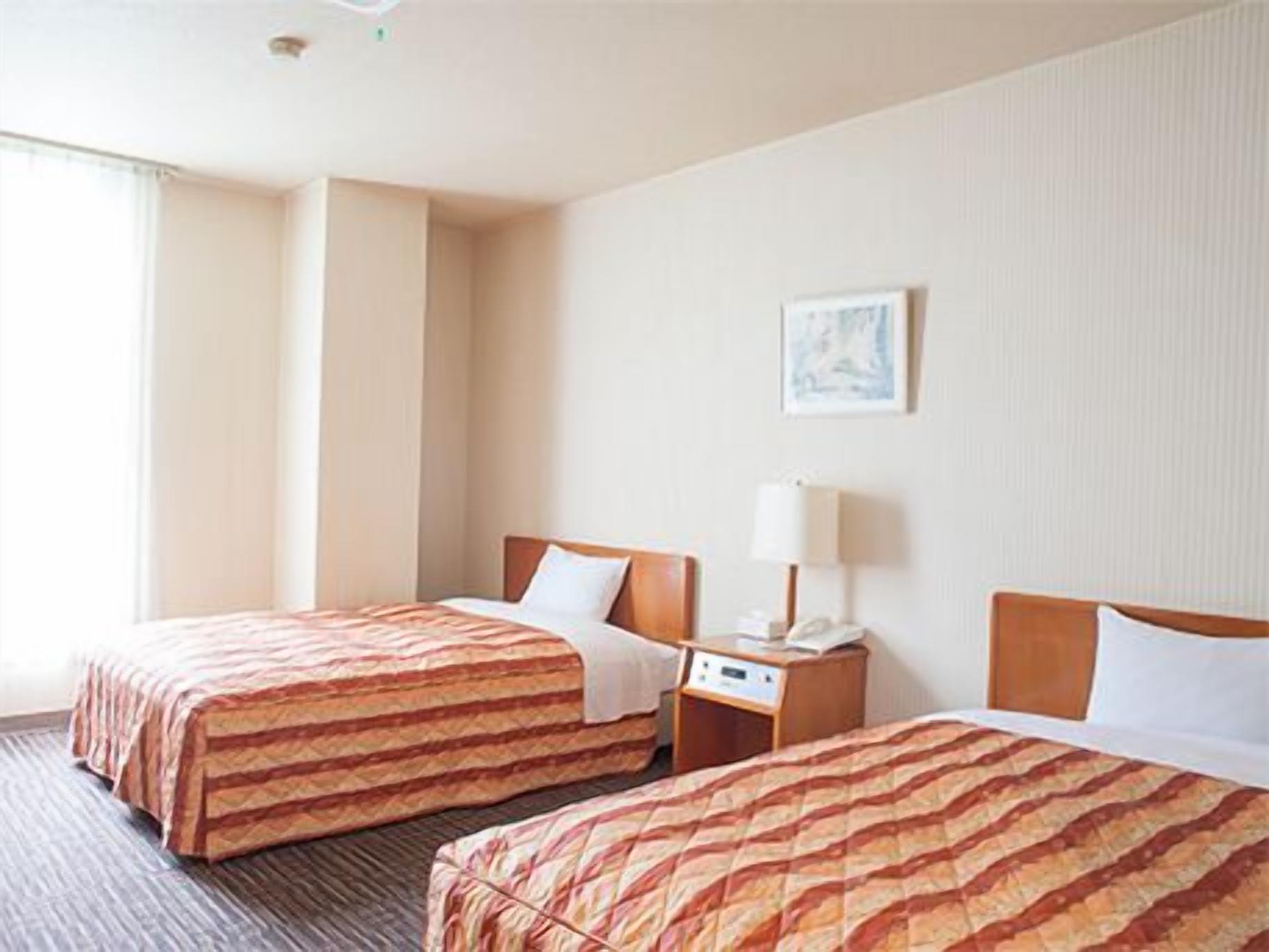 A hotel room with two beds, a bedside table with a lamp, a painting on the wall, and a window.