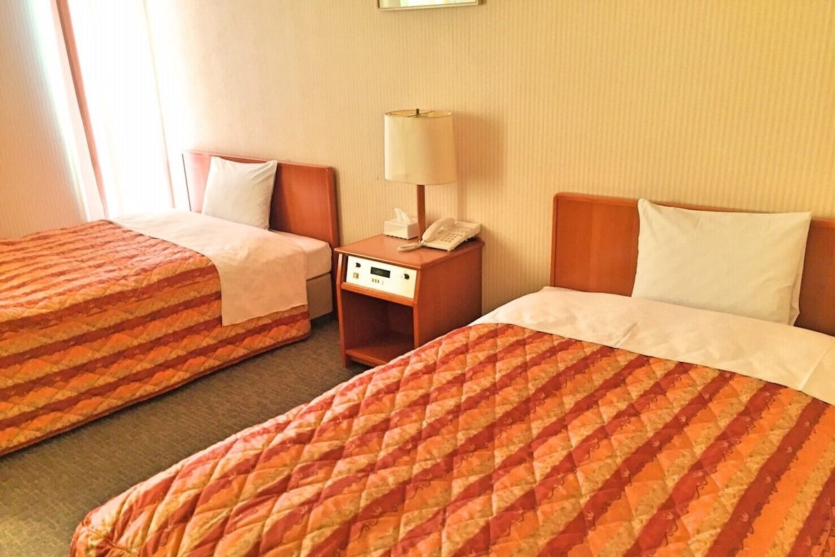 Habitación de hotel con dos camas, cada una con colcha y almohadas estampadas, una mesita de noche con lámpara y teléfono, y una lámpara de pared.