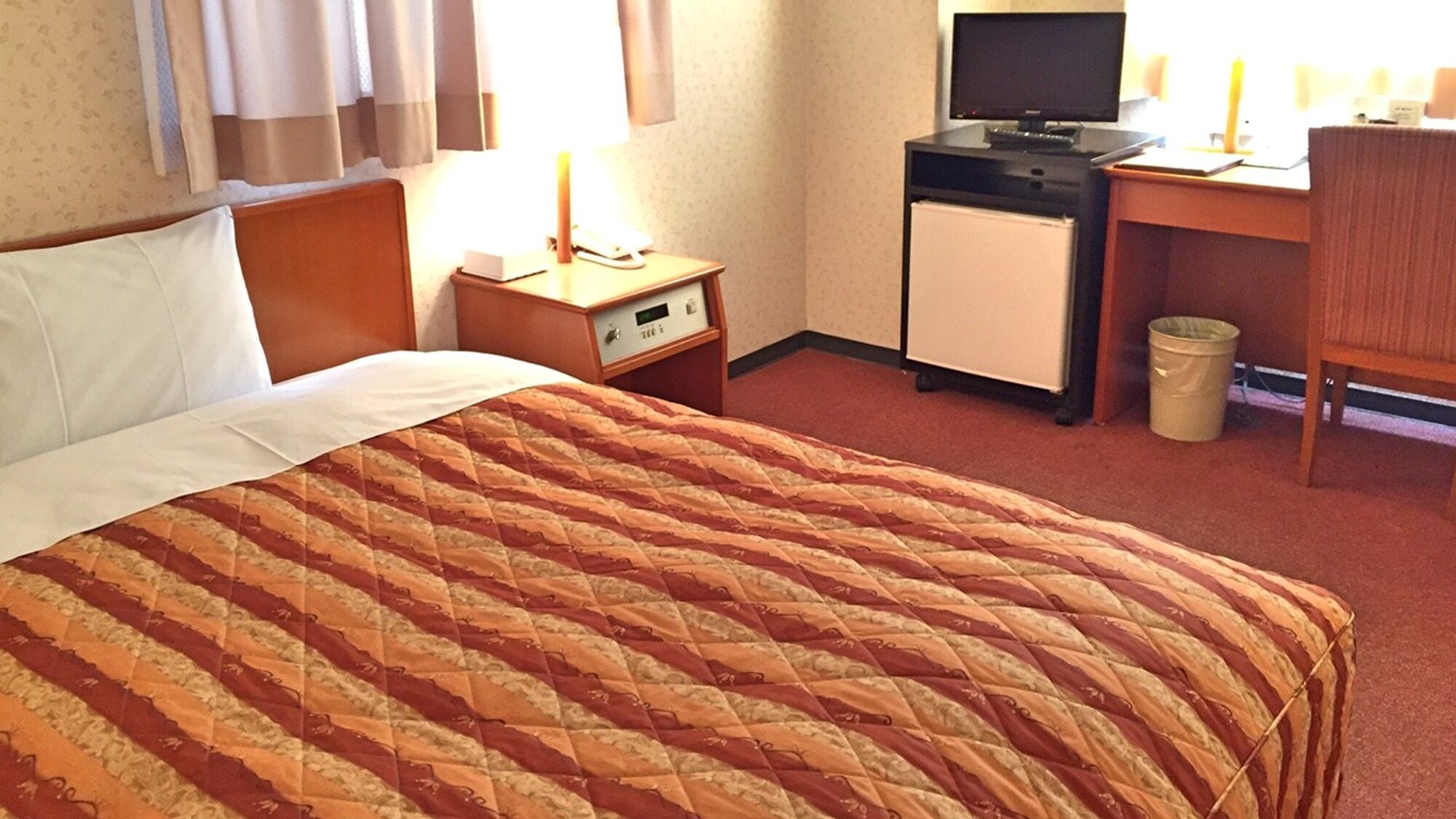 A hotel room with a bed, a desk with a computer, a small refrigerator, and a trash bin.