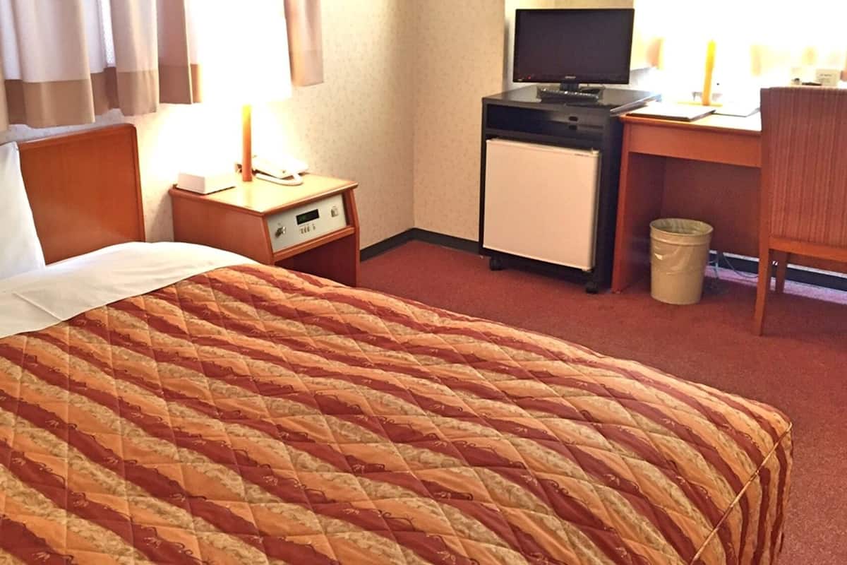 Habitación de hotel con cama, escritorio con computadora, refrigerador pequeño y cesto de basura.