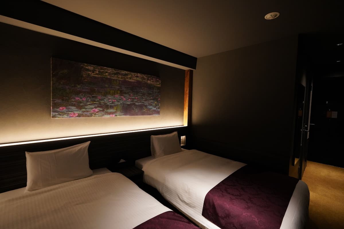 Quarto de hotel com duas camas, uma pintura de nenúfares e uma cabeceira escura.