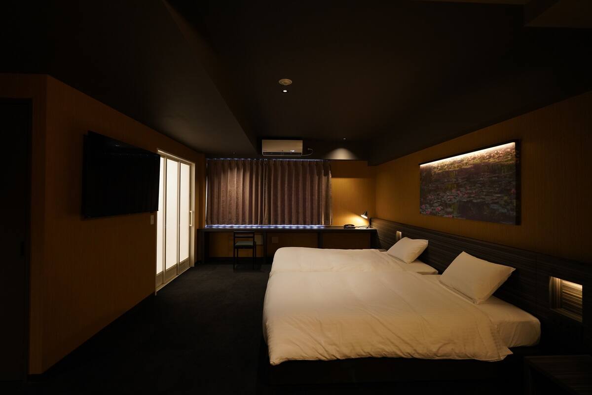 Quarto de hotel com uma cama grande, escrivaninha, cadeira, televisão e um quadro na parede.