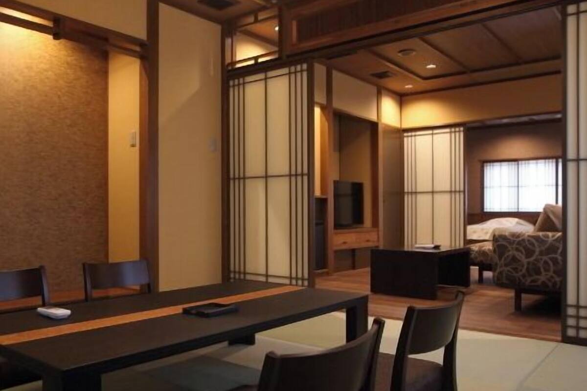 Um quarto tradicional em estilo japonês, com tatames, uma mesa baixa com cadeiras, uma cama com colcha estampada e portas corrediças.