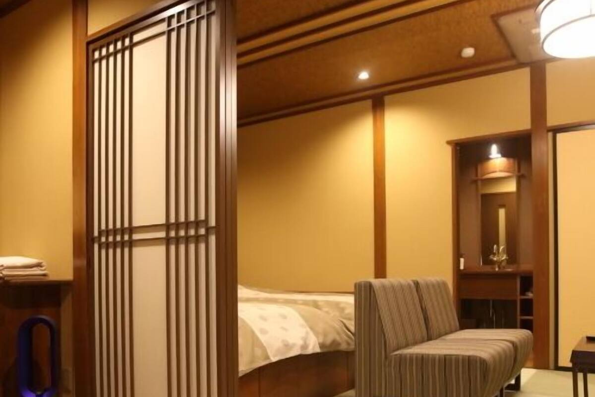 Quarto com cama, cadeira, mesa pequena e um separador com ripas verticais.