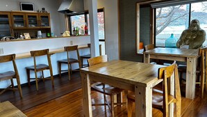 A wooden dining area with chairs and a table, a bar with bottles, and a large window with a view. - Nora (Hokuto)