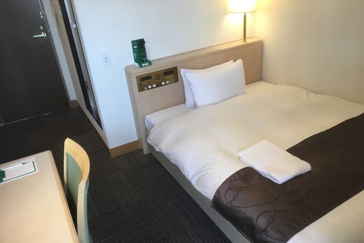Una habitación de hotel con una cama, un escritorio, una silla, una mesita de noche y una lámpara.