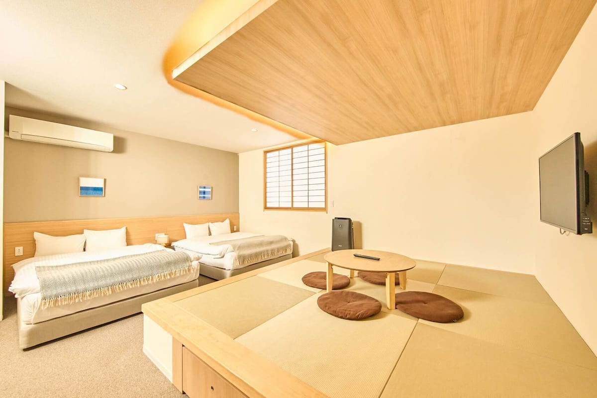 A hotel room with two beds, a small table with cushions, a TV, and a window with blinds.
