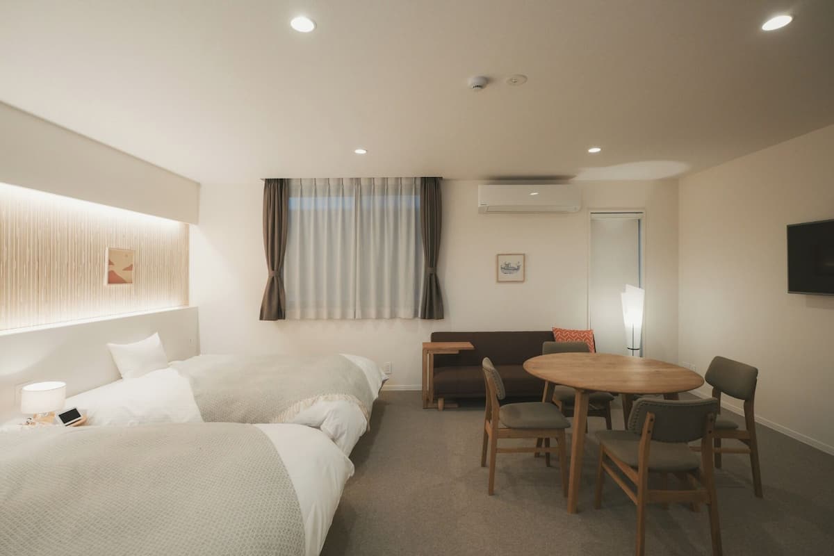 A hotel room with two beds, a dining table with chairs, a flat-screen TV, and an air conditioning unit.