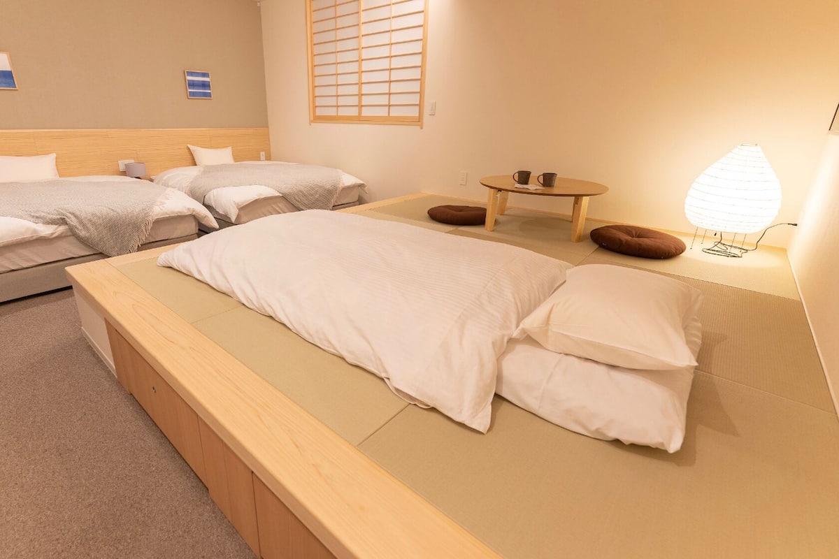 A hotel room with two single beds, a sofa bed, a small table with two cups, a lamp, and a window with a view.