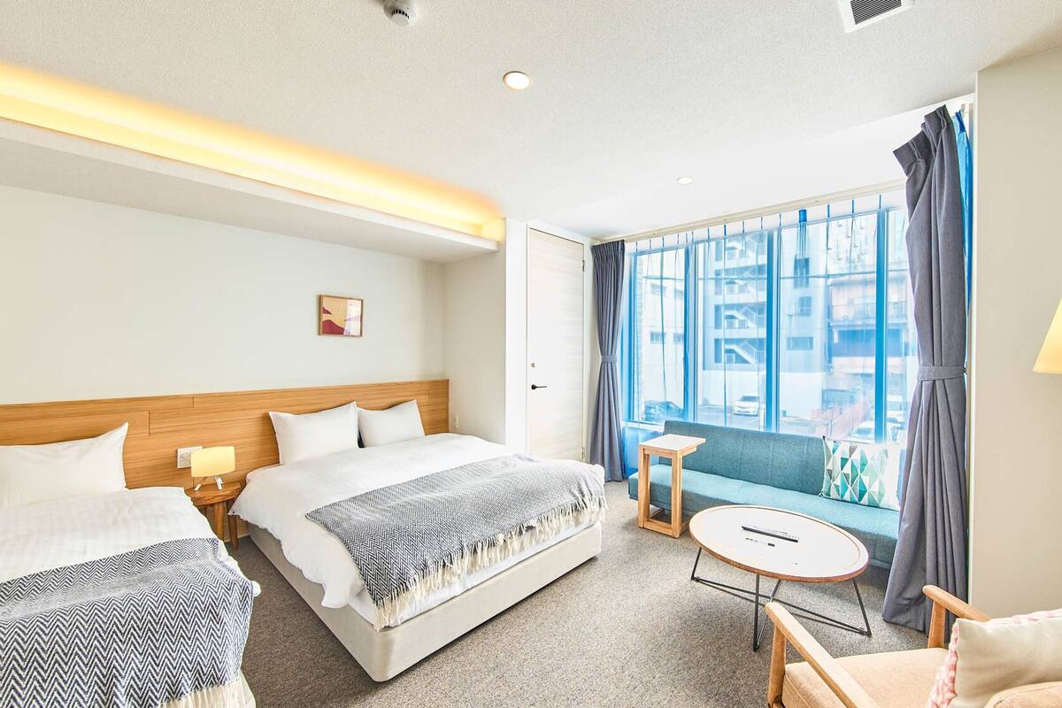 A hotel room with two beds, a sofa, a small table, and a view of the city through large windows.