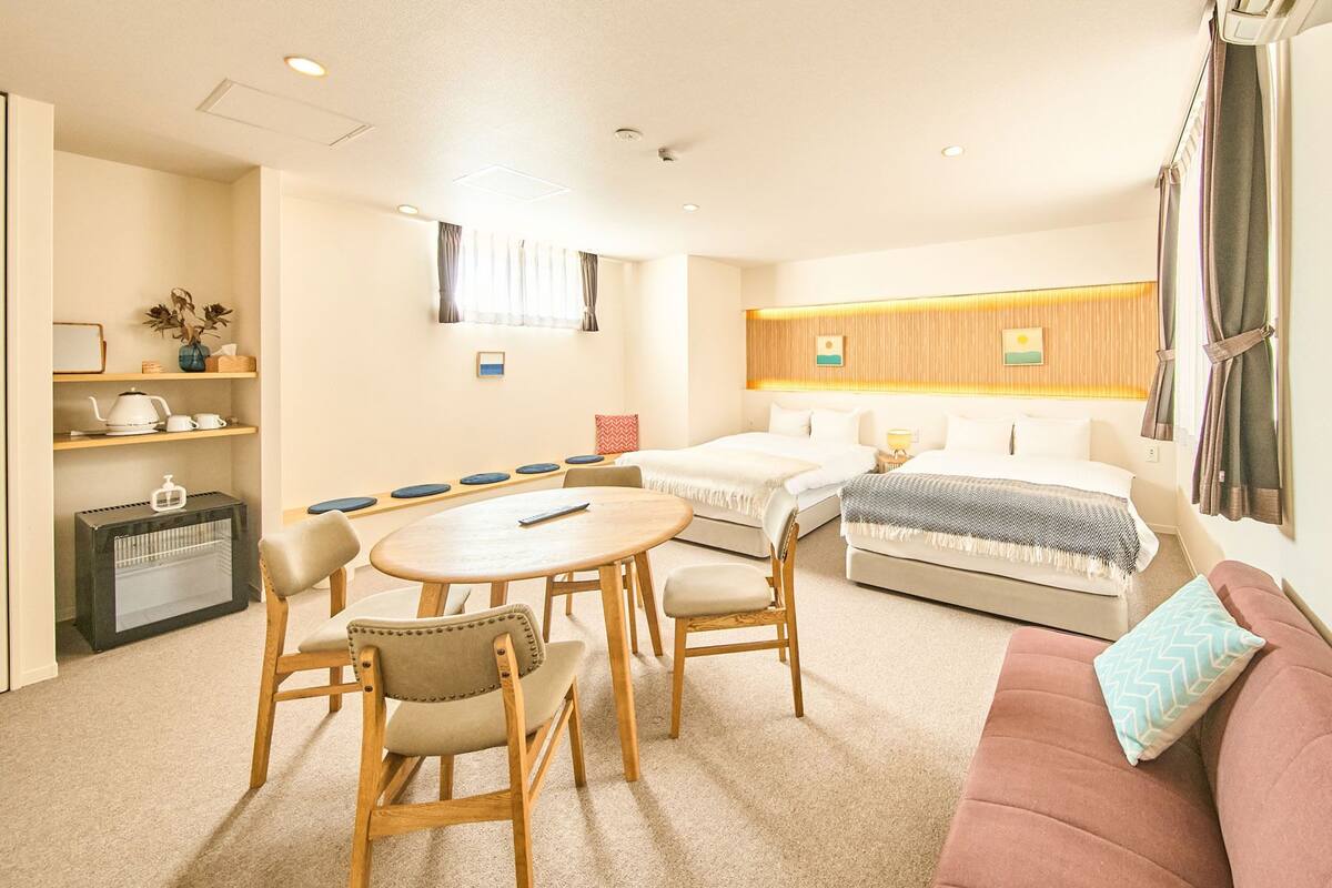 A hotel room with a sofa, a round table with chairs, a bed, a small table with a lamp, and a shelf with items.