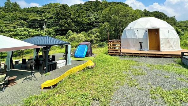 A tent with a wooden deck, a small table, and a yellow slide in a grassy area with a forested hill in the background. - Nora (Hokuto)