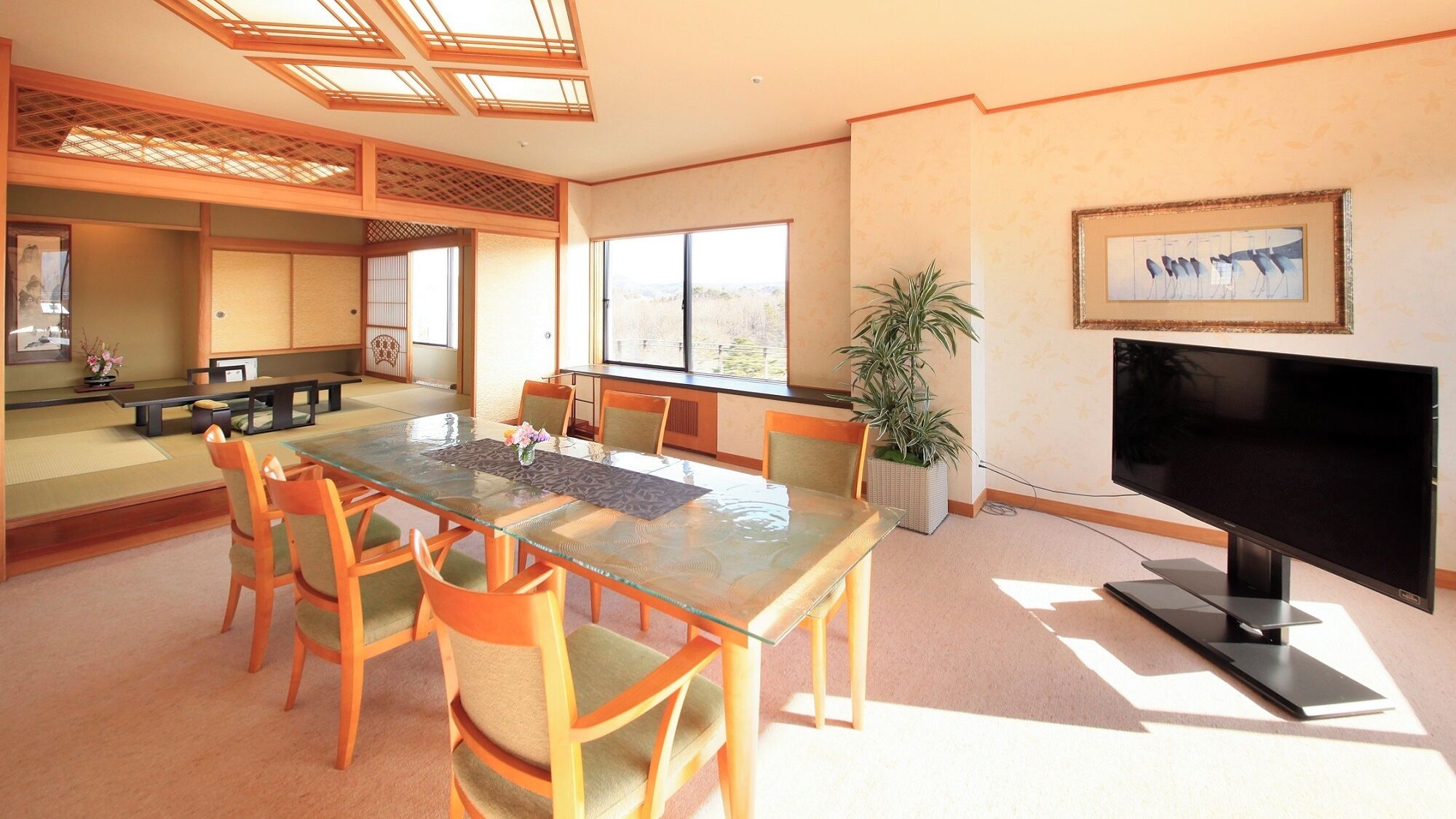 A traditional Japanese-style room with tatami flooring, a low table with cushions, a television, and a large window.