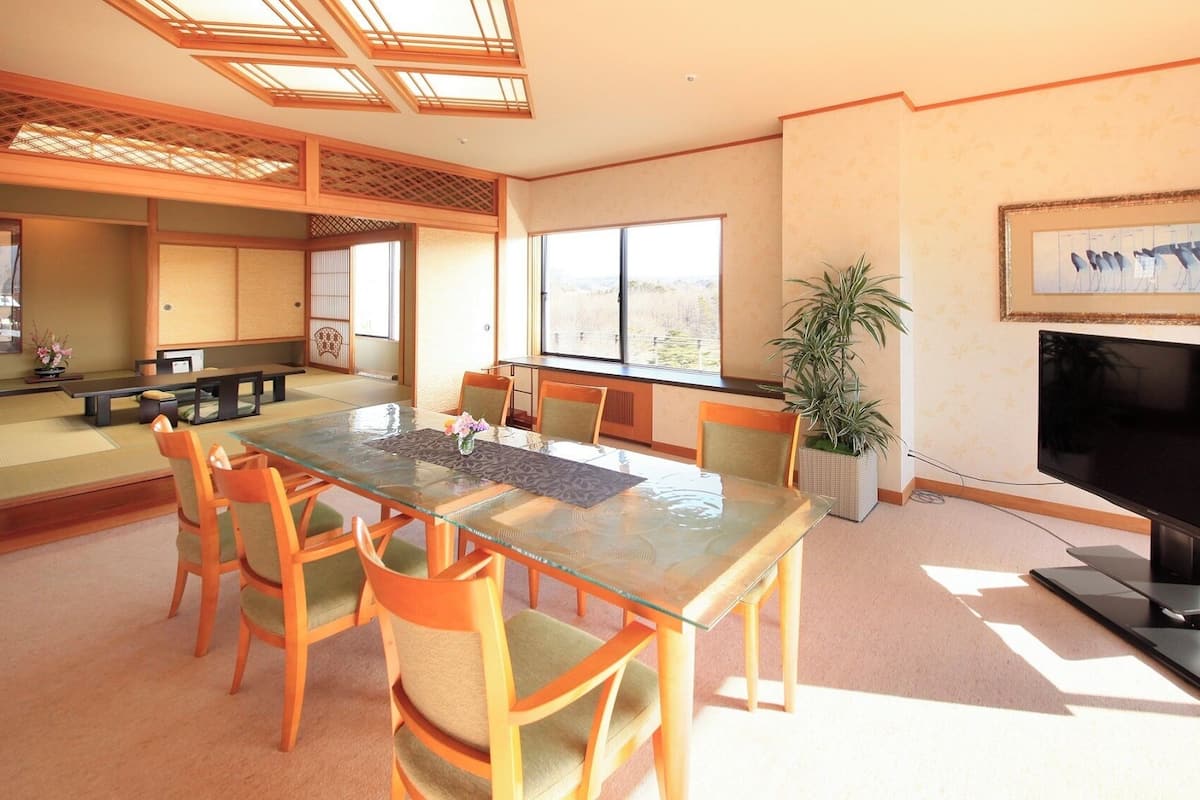 Una habitación tradicional japonesa con suelo de tatami, una mesa baja con cojines, televisor y un ventanal grande.