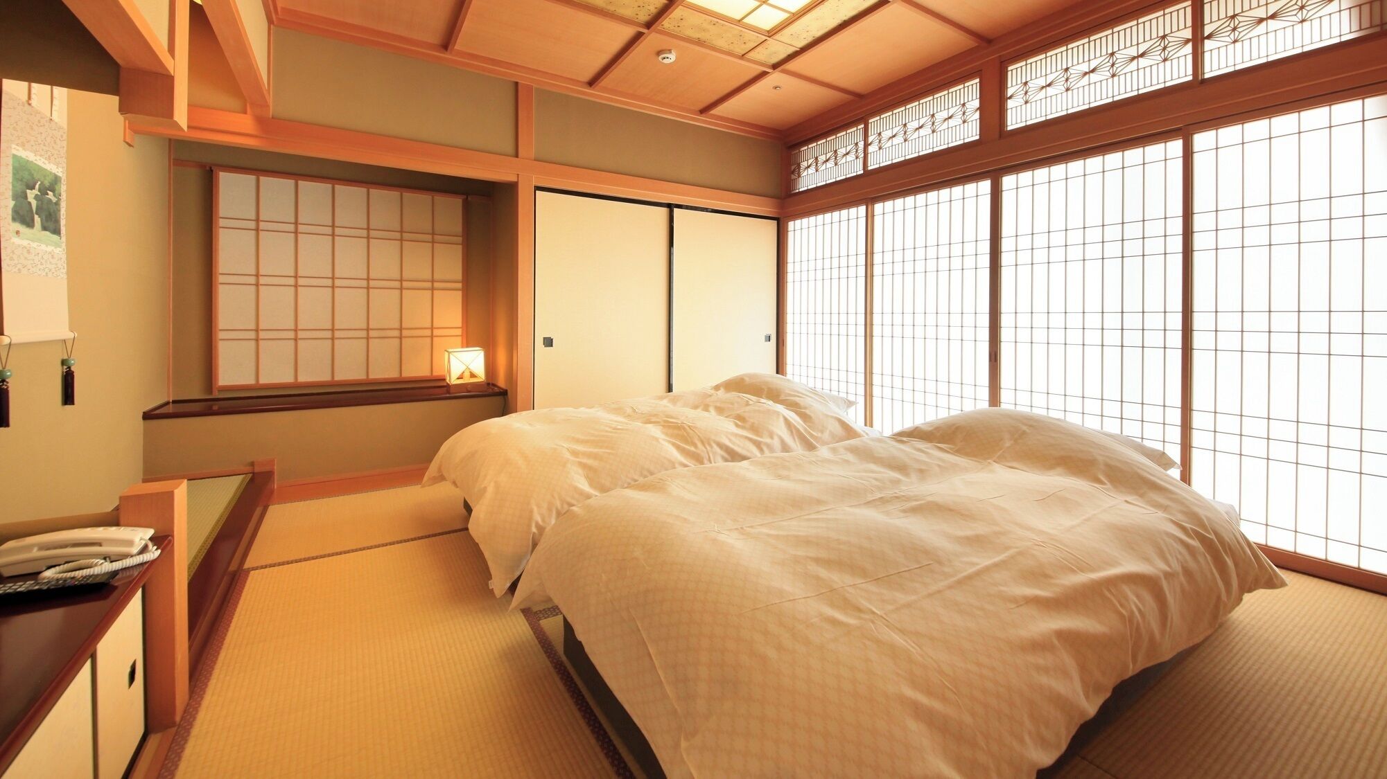 A traditional Japanese-style room with tatami flooring, a bed with white bedding, a desk with a telephone, and sliding doors.