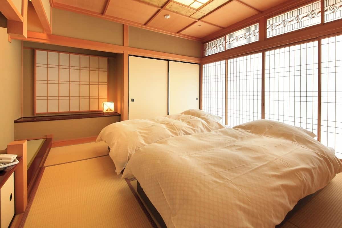 Una habitación tradicional japonesa con suelo de tatami, una cama con ropa de cama blanca, un escritorio con teléfono y puertas correderas.