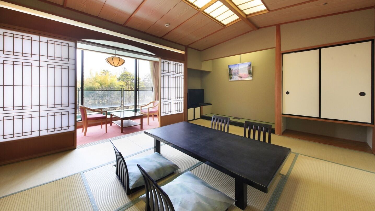 A traditional Japanese room with tatami flooring, a low table, chairs, a sliding door, and a view through a large window.