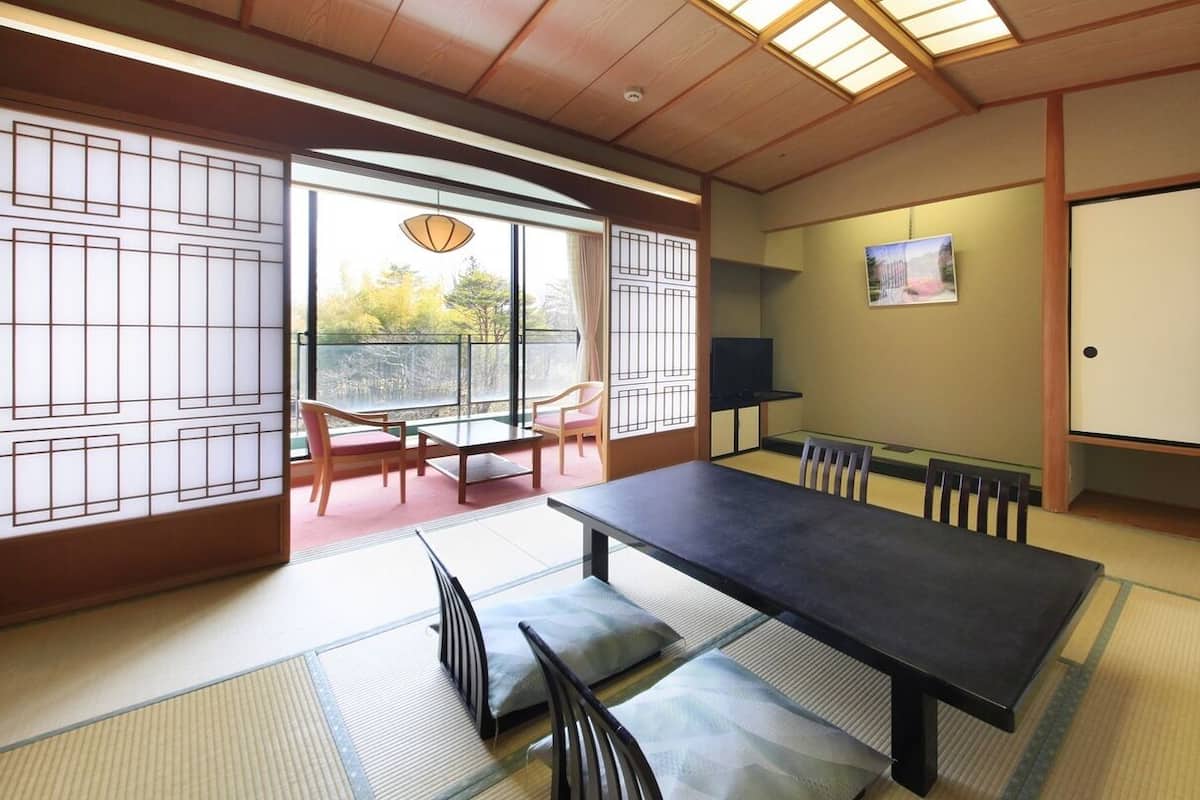 Una habitación japonesa tradicional con suelo de tatami, una mesa baja, sillas, una puerta corredera y vistas a través de un ventanal.
