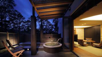 Un jacuzzi en un área de terraza de madera con una pérgola arriba, un espacio de estar con una mesa y sillas, y vistas a árboles al anochecer.