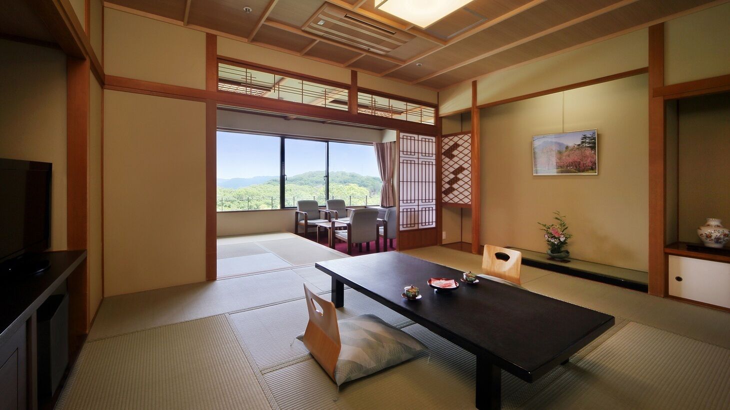 Una habitación japonesa tradicional con suelo de tatami, una mesa baja, sillas, televisor y vistas al exterior.