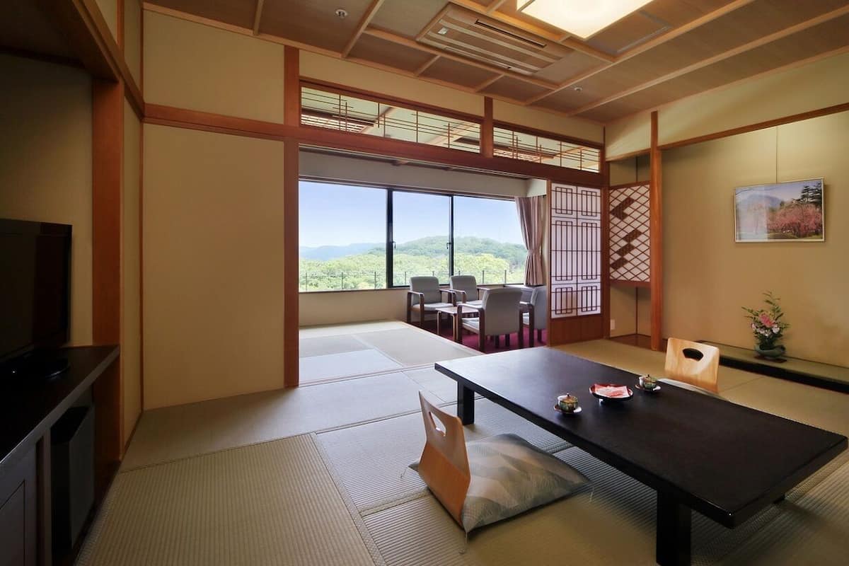 Una habitación japonesa tradicional con suelo de tatami, una mesa baja, sillas, televisor y vistas al exterior.
