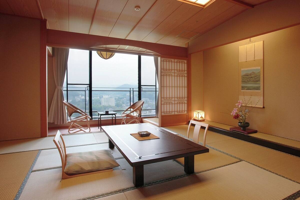 Una habitación tradicional japonesa con suelo de tatami, una mesa baja y vistas a un paisaje urbano a través de grandes ventanales.